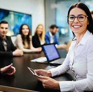 planning_meeting_woman_smile.jpg A woman holding a tablet looks into the camera while sitting at a conference table during a business meeting